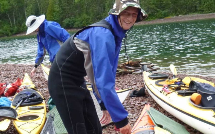 A person smiles at the camera while tending to kayaks resting on the shore of a lake. A person does the same in the background. 